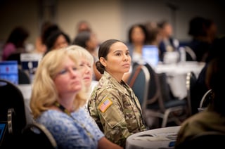 Woman in camouflage sits in a room surrounded by other people. 