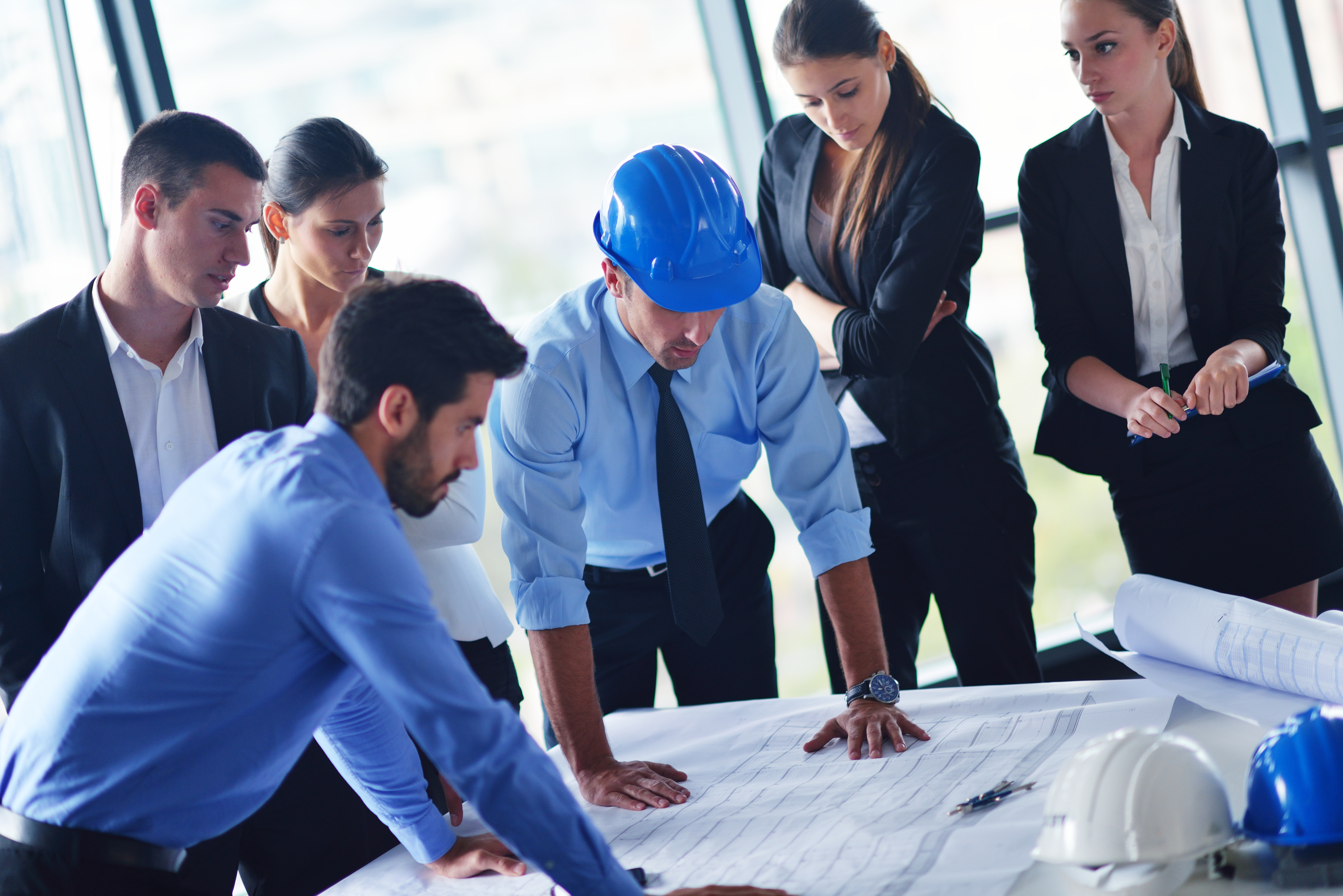 Six people stand and look at blueprints on a table. One man is wearing a hard hat.