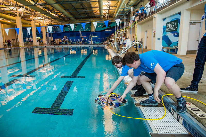 Two teen boys lower a remote operated vehicle into a swimming pool for robotics competition.